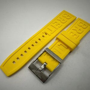 Puede incluir: Correa de reloj Breitling de color amarillo brillante con hebilla plateada. La correa est&aacute; hecha de un material flexible y presenta el nombre de la marca en relieve. La hebilla tiene el logotipo de Breitling.