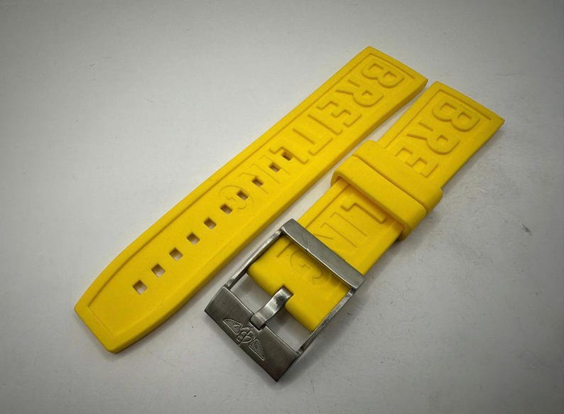 Puede incluir: Correa de reloj Breitling de color amarillo brillante con hebilla plateada. La correa est&aacute; hecha de un material flexible y presenta el nombre de la marca en relieve. La hebilla tiene el logotipo de Breitling.