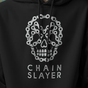 Puede incluir: Sudadera negra con un diseño de calavera blanca hecho con cadenas de bicicleta. El texto "CHAIN SLAYER" está impreso debajo de la calavera. La sudadera tiene un bolsillo delantero y una capucha.