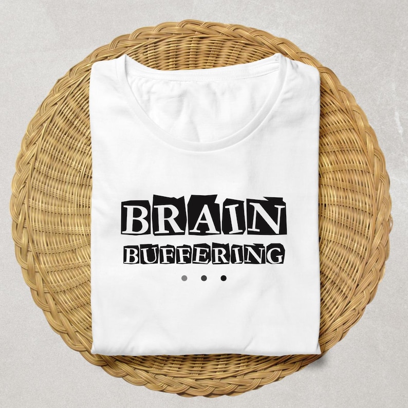 May include: A white short-sleeved t-shirt featuring the black text "BRAIN BUFFERING" printed in a bold, blocky font across the chest, accompanied by three small black dots arranged horizontally underneath the text to represent a loading icon.