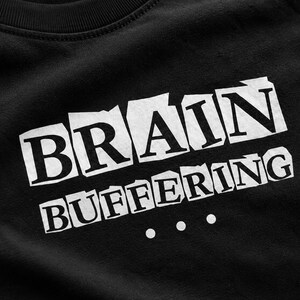 May include: A black t-shirt featuring the white text BRAIN BUFFERING with three dots below it. The letters are presented in a ransom note style with individual white rectangular backgrounds for each letter, creating a high-contrast graphic design.