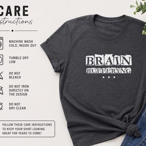 May include: A dark charcoal gray t-shirt featuring the white text BRAIN BUFFERING with three dots below. To the left, a care instruction chart lists machine wash cold, tumble dry low, do not bleach, do not iron on design, and do not dry clean.