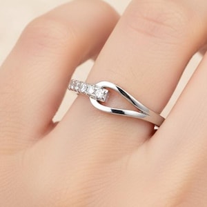 Puede incluir: Un anillo de plata con un diseño único. El anillo presenta una fila de pequeñas piedras transparentes engastadas en una banda curva, con un detalle de bucle. El anillo se muestra en un dedo.