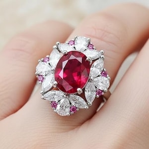 Peut inclure: Une bague en argent ornée d'un grand rubis central de forme ovale. Le rubis est entouré d'un halo de pierres blanches et roses, créant un motif floral. La bague est montée sur un anneau en argent.