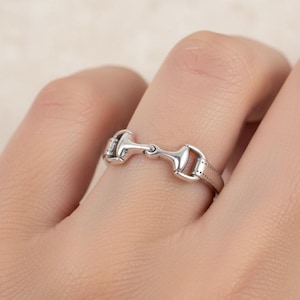 May include: A silver-colored ring in the shape of a horse bit. The ring is worn on a finger and has a detailed design with a polished finish. The ring is a piece of equestrian-themed jewelry.