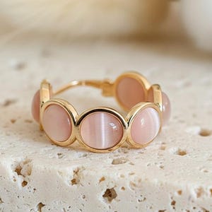 Può includere: Un anello d'oro con pietre preziose ovali rosa chiaro. Le pietre sono disposte in un motivo ripetuto attorno all'anello. L'anello è esposto su una superficie chiara e strutturata.