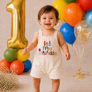 Può includere: Una tutina senza maniche color crema con il testo ricamato "It's my birthday" in lettere multicolori, accompagnato da piccole icone ricamate di una torta di compleanno, palloncini e regali sulla zona del petto.