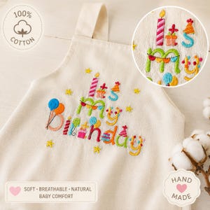 Può includere: Una tutina per neonati in cotone bianco sporco con testo ricamato colorato che recita It's My Birthday. Il design include motivi festivi come candele, cappellini da festa, palloncini e stelle. Un inserto circolare mostra un primo piano del ricamo.