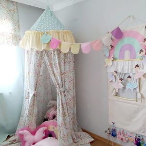 May include: A floral print canopy tent with a light blue polka dot top and a yellow frill. A garland of pastel-coloured flags hangs nearby. Pink cushions and a rainbow-shaped cushion are inside the tent, creating a whimsical play space.