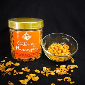 Puede incluir: Un tarro transparente con tapa dorada y etiqueta naranja con la leyenda "Catering Handayani" contiene cebollas fritas. Un pequeño cuenco de cristal también contiene cebollas fritas, con más esparcidas sobre una superficie negra.