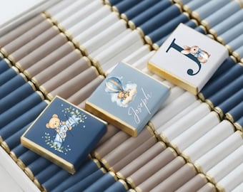 May include: A box of chocolates with a variety of colors, including blue, white, and brown. Some chocolates have a printed design with a teddy bear, hot air balloon, and the letter "J". The chocolates are arranged in rows.