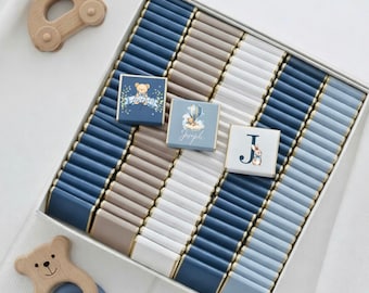 May include: A white box filled with rows of blue, gray, and white chocolate bars. Three decorated chocolates are visible, one with a teddy bear, one with a hot air balloon, and one with the letter "J" and a bunny. Wooden baby toys are also present.