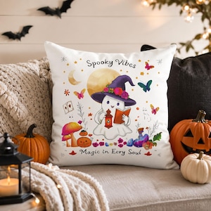 May include: A white square throw pillow featuring a cartoon ghost wearing a purple witch hat with flowers, holding a lantern and an open book. The text reads Spooky Vibes and Magic in Eery Soul, surrounded by moons, stars, butterflies, and mushrooms.