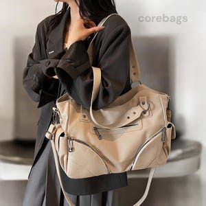 Peut inclure: Un sac à main en cuir beige avec plusieurs compartiments zippés et une bandoulière. Le sac a un design structuré avec des accessoires argentés et une poignée supérieure. Le mot "corebags" est visible dans le coin supérieur droit.