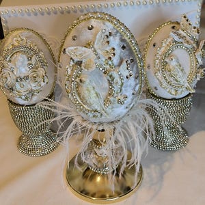 Op de afbeelding: Drie decoratieve eivormige ornamenten in wit en goud, versierd met strasssteentjes, parels en reliëffiguren van vogels, vlinders en rozen. Elk ei rust op een goudkleurige voet die bedekt is met kleine, fonkelende kristallen.