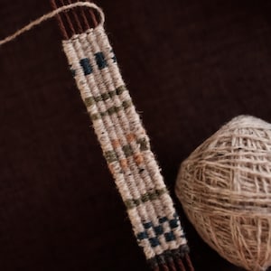 May include: A close-up of a weaving project in progress. The woven piece is a narrow strip with a geometric pattern in shades of white, blue, green, and brown. A ball of yarn sits next to the woven strip. The background is a solid brown.