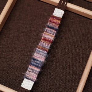 Puede incluir: Primer plano de un pequeño textil tejido y colorido sobre un telar de madera. La pieza tejida presenta una mezcla de colores, incluyendo rosa, azul, naranja y blanco. El telar es de madera clara y está sobre un fondo marrón.