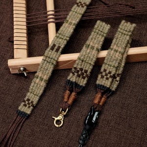 May include: Handwoven textile straps in shades of green, brown, and black. The straps feature a geometric pattern and are attached to a wooden frame. One strap has a gold-colored clasp, and another has a black leather loop.