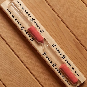 Può includere: Una clessidra di vetro trasparente con sabbia rossa, incastonata in una cornice di legno chiaro. La cornice presenta segni neri che indicano incrementi di tempo di 5, 10 e 15 minuti. La clessidra è posizionata in diagonale.