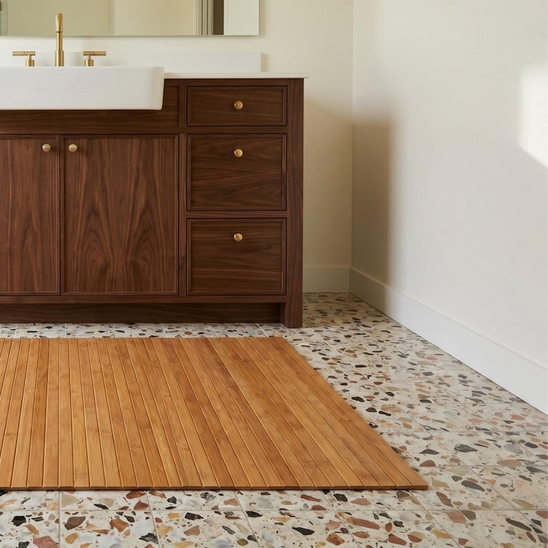 K&ouml;nnte beinhalten: Eine hellbraune Badematte aus Holz mit Lamellendesign liegt auf einem gefleckten Fliesenboden. Die Matte befindet sich vor einem dunklen Holzwaschtisch mit goldfarbener Hardware und einem wei&szlig;en Waschbecken. &Uuml;ber dem Waschbecken befindet sich ein Spiegel.