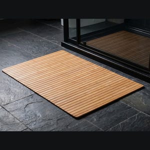 K&ouml;nnte beinhalten: Eine rechteckige, hellbraune Badematte aus Holz mit Lamellendesign. Die Matte liegt auf einem dunkelgrauen Fliesenboden vor einer schwarz gerahmten Duschkabine. Das Holz hat eine nat&uuml;rliche Maserung.