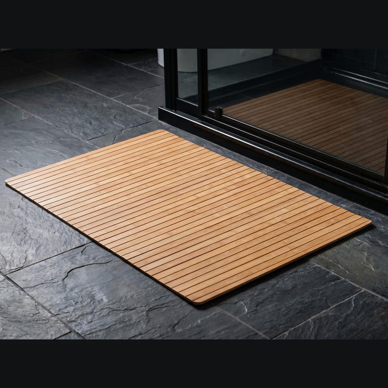 K&ouml;nnte beinhalten: Eine rechteckige, hellbraune Badematte aus Holz mit Lamellendesign. Die Matte liegt auf einem dunkelgrauen Fliesenboden vor einer schwarz gerahmten Duschkabine. Das Holz hat eine nat&uuml;rliche Maserung.