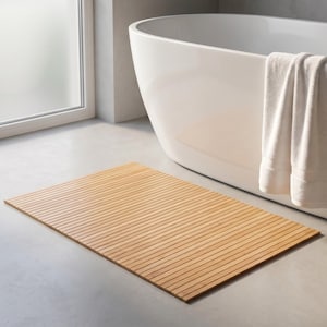 K&ouml;nnte beinhalten: Eine rechteckige, hellbraune Badematte aus Holz mit Lamellendesign. Die Matte liegt auf einem grauen Betonboden in der N&auml;he einer wei&szlig;en Badewanne und eines Fensters. Ein wei&szlig;es Handtuch h&auml;ngt an der Seite der Wanne.