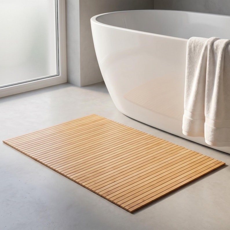 K&ouml;nnte beinhalten: Eine rechteckige, hellbraune Badematte aus Holz mit Lamellendesign. Die Matte liegt auf einem grauen Betonboden in der N&auml;he einer wei&szlig;en Badewanne und eines Fensters. Ein wei&szlig;es Handtuch h&auml;ngt an der Seite der Wanne.