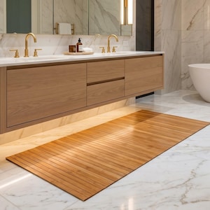 K&ouml;nnte beinhalten: Ein rechteckiger Badteppich aus nat&uuml;rlichem Bambus mit Lamellendesign. Der hellbraune Teppich liegt auf einem wei&szlig;en Marmorboden in einem modernen Badezimmer. Das Badezimmer verf&uuml;gt &uuml;ber einen Holzwaschtisch, goldfarbene Armaturen und eine wei&szlig;e Badewanne.