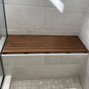 May include: A rectangular shower bench seat constructed from horizontal dark brown wooden slats. The bench features a slatted design with small gaps between each board, resting flat on a white tiled ledge within a tiled shower enclosure.