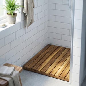 May include: A wooden shower mat made of parallel wooden slats. The mat is rectangular and fits inside a shower stall with white tiled walls. A small potted plant sits on a windowsill above the mat.