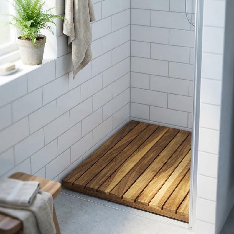 May include: A wooden shower mat made of parallel wooden slats. The mat is rectangular and fits inside a shower stall with white tiled walls. A small potted plant sits on a windowsill above the mat.