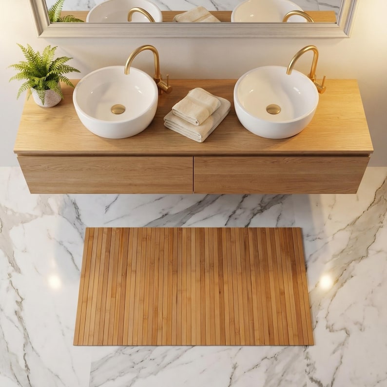 K&ouml;nnte beinhalten: Ein moderner Badezimmerwaschtisch mit zwei wei&szlig;en, runden Waschbecken und goldenen Wasserh&auml;hnen. Der Waschtisch ist aus hellem Holz gefertigt und hat zwei Schubladen. Eine Bambus-Badematte liegt auf dem Marmorboden. Eine kleine Pflanze und gefaltete Handt&uuml;cher sind ebenfalls zu sehen.