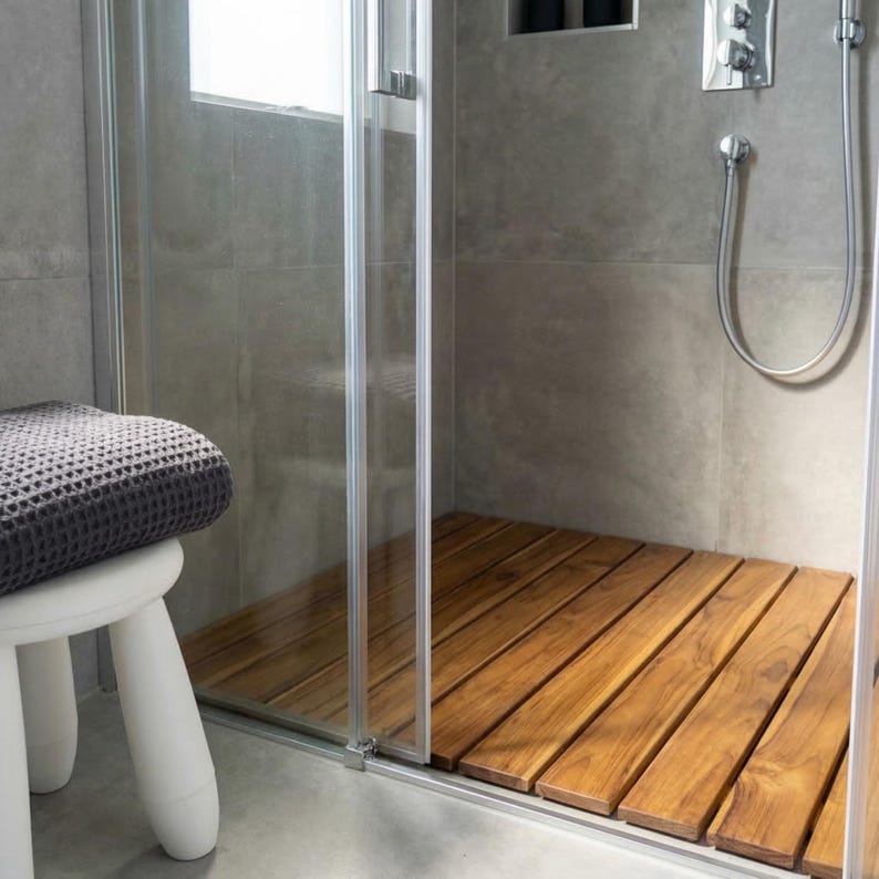 May include: A modern shower stall with a wooden floor made of rectangular planks. The shower has a glass door and gray tiled walls. A small white stool with a gray towel is visible to the left.