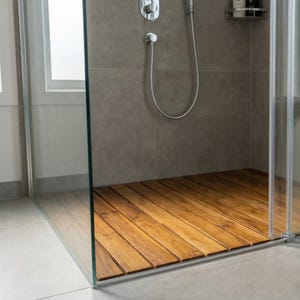 May include: A modern shower enclosure with a wooden floor. The floor is made of light brown wooden planks. The shower has a glass door and a silver shower head and faucet. The walls are a neutral gray color.