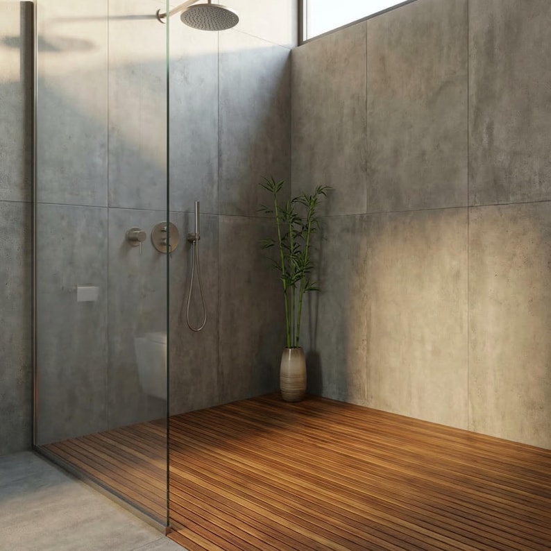 May include: Modern shower with gray concrete walls, a glass enclosure, and a wooden floor. A silver shower head and fixtures are mounted on the wall. A vase with green plants sits in the corner, adding a touch of nature to the space.