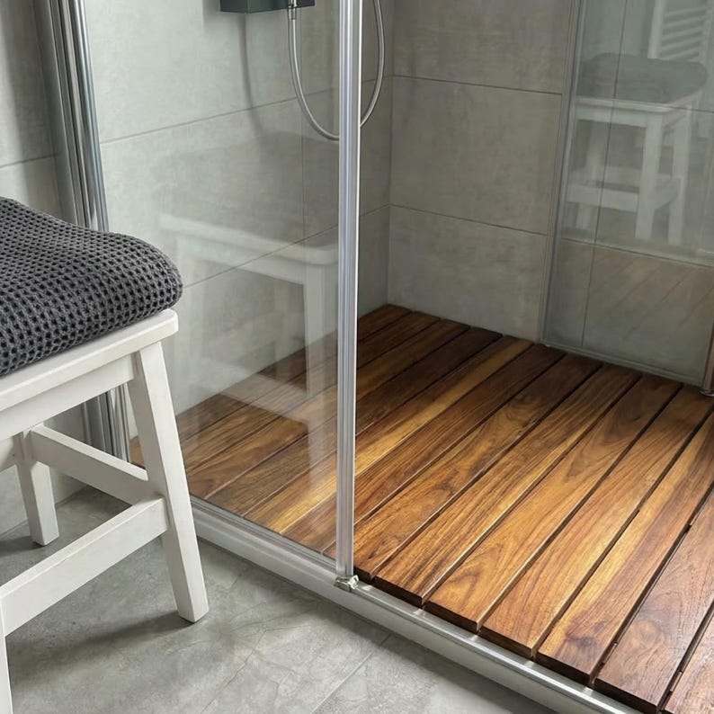 May include: A wooden shower floor with a natural wood grain pattern. The shower floor is inside a glass enclosure. A white stool with a gray towel is in the foreground.