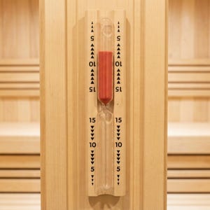 May include: A clear glass hourglass filled with red sand, mounted on a light wood panel. The panel has markings for 5, 10, and 15 minutes. The hourglass is designed for use in a sauna or steam room.