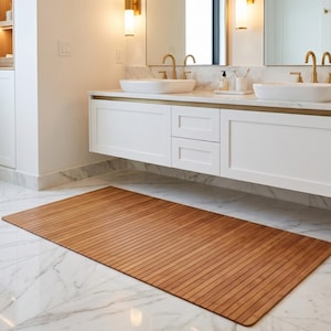 K&ouml;nnte beinhalten: Eine rechteckige Badematte aus Holz mit Lamellendesign. Die Matte besteht aus hellbraunem Holz und liegt auf einem wei&szlig;en Marmorboden in einem Badezimmer. Das Badezimmer verf&uuml;gt &uuml;ber wei&szlig;e Schr&auml;nke, goldfarbene Armaturen und einen gro&szlig;en Spiegel.