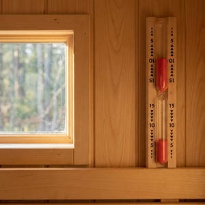 May include: A wooden sauna interior with a window and a red hourglass timer. The timer is marked with 5, 10, and 15-minute increments. The sauna walls and trim are made of light-colored wood.