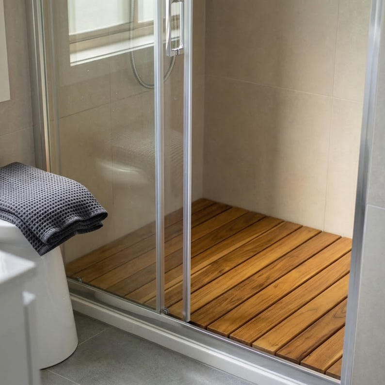 May include: A modern shower stall with a clear glass door and a wooden floor. The floor is made of long, narrow planks of light brown wood. A gray waffle-weave towel rests on a white fixture to the left of the shower.