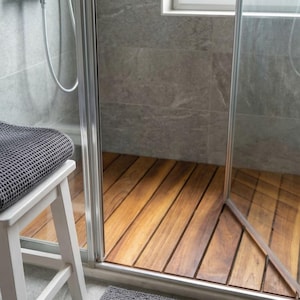 May include: A modern shower with a wooden floor and glass door. A gray towel rests on a white stool. The shower walls are gray tile, and a window is visible in the background. The wooden floor is made of long, narrow planks.
