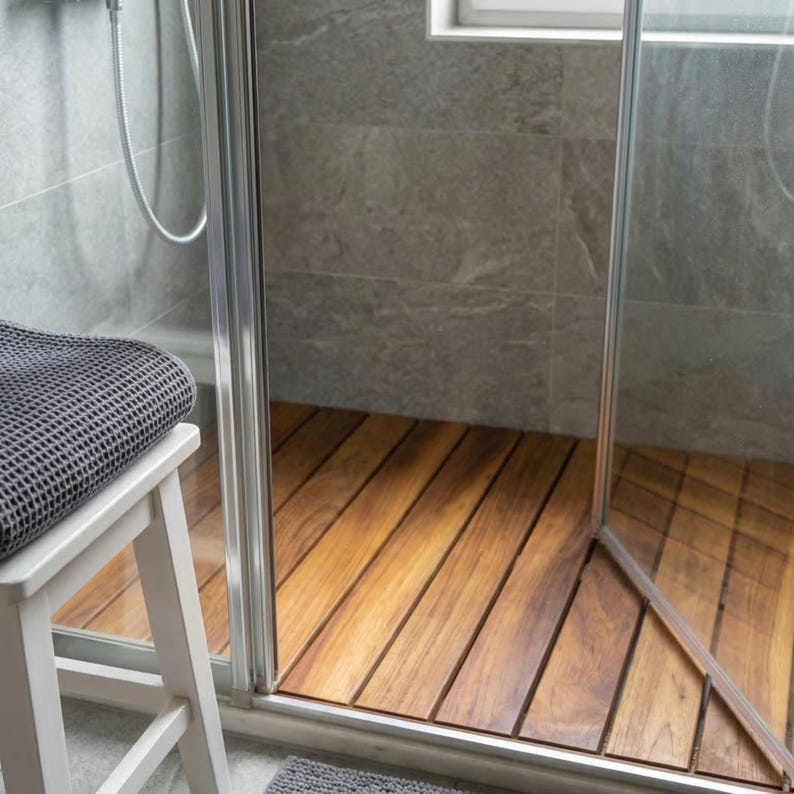 May include: A modern shower with a wooden floor and glass door. A gray towel rests on a white stool. The shower walls are gray tile, and a window is visible in the background. The wooden floor is made of long, narrow planks.