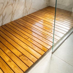 Può includere: Un tappetino per doccia in legno composto da doghe di legno parallele, marrone chiaro. Il tappetino è bagnato, con goccioline d'acqua visibili sulla superficie. Il tappetino è posto all'interno di un box doccia con porta in vetro e parete marmorizzata.