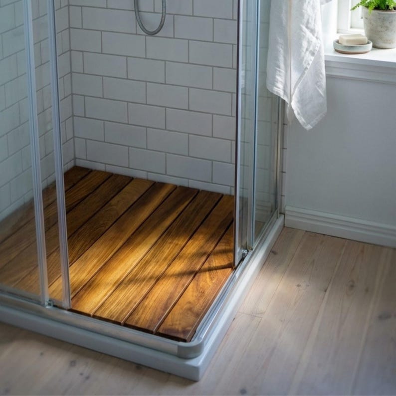 May include: A shower stall with a wooden floor made of planks. The shower has glass doors and white tile walls. A white towel hangs on the outside of the shower. The wooden floor is a rich brown color.