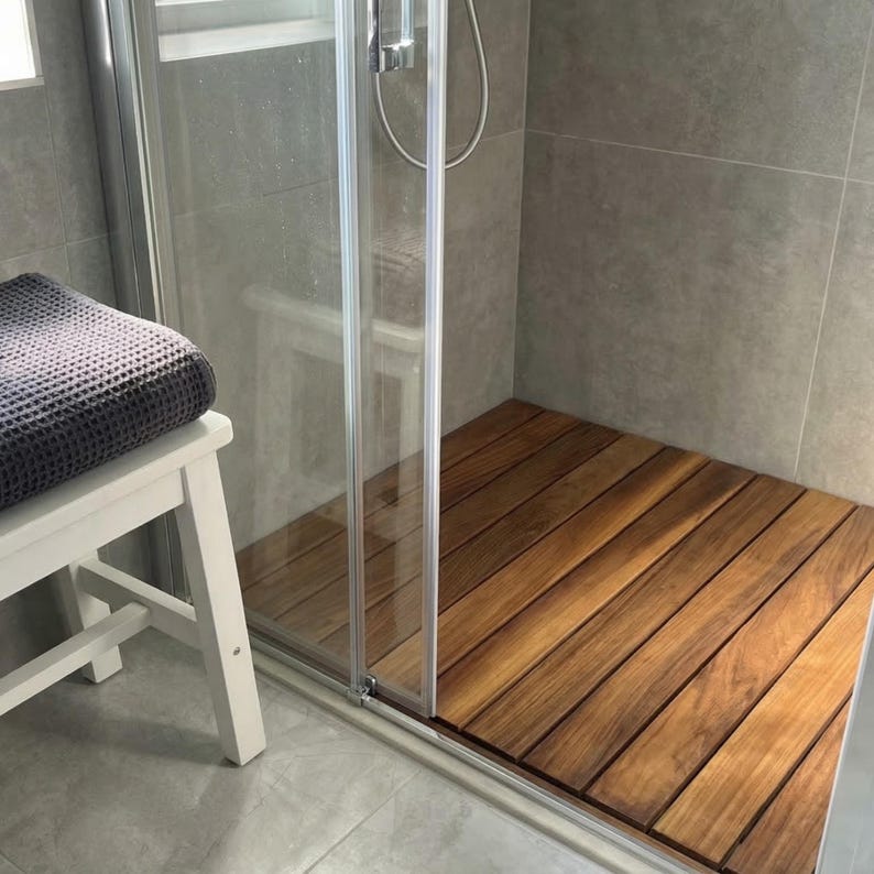 May include: A wooden shower floor made of planks of wood. The shower has a glass door and is next to a white bench with a gray towel. The shower walls are gray tile.