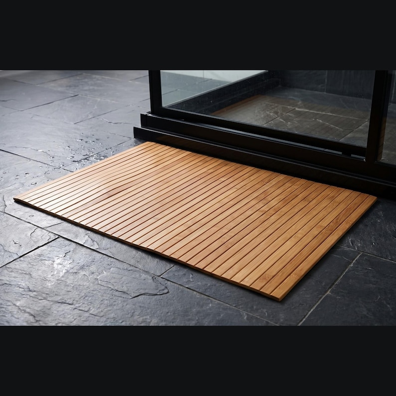 K&ouml;nnte beinhalten: Eine rechteckige Badematte aus Holz mit Lamellendesign. Die Matte besteht aus hellbraunem Holz und wird vor einer Glas-Duschabt&uuml;r platziert. Die Matte ist ca. 61 cm lang und 41 cm breit.