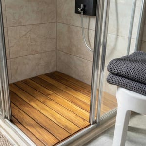 May include: A shower stall with a wooden floor made of parallel planks. The stall has a glass door and a black shower head. A stack of gray towels rests on a white stool outside the shower.