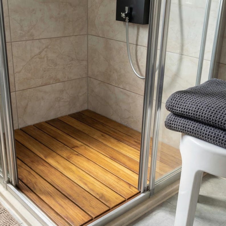 May include: A shower stall with a wooden floor made of parallel planks. The stall has a glass door and a black shower head. A stack of gray towels rests on a white stool outside the shower.
