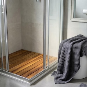 May include: A modern shower enclosure with a wooden floor. The shower has a glass door and a silver frame. A gray waffle-weave towel is draped over a white stool, suggesting a spa-like bathroom setting.